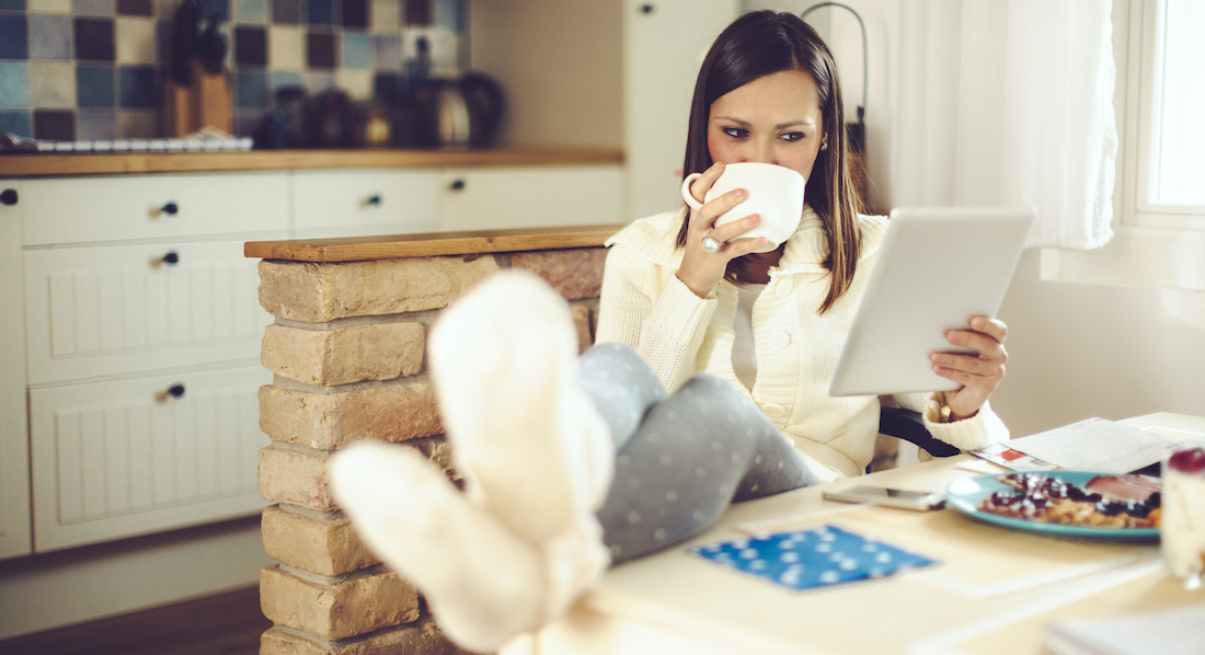 Eine junge Frau sitzt entspannt bei einer Tasse Kaffee in der Küche und nutzt auf ihrem Tablet ein TAN-Verfahren für den Online-Einkauf