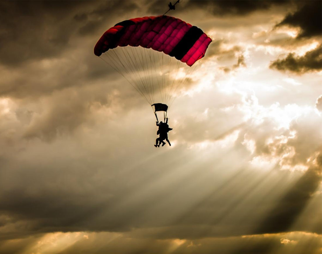 Ein Fallschirm schwebt vor dem Hintergrund düsterer Wolken, durch die einzelne Lichtstrahlen brechen, herab