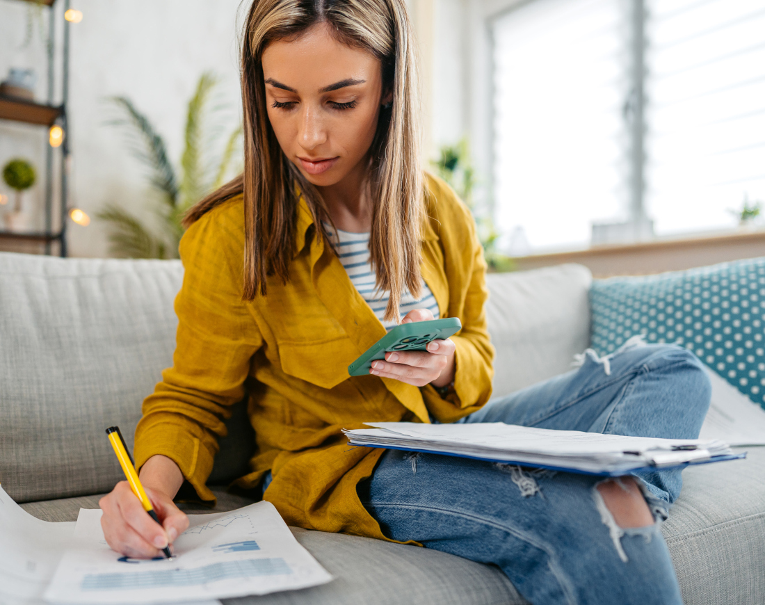 Eine junge Frau sitzt mit ihrem Handy und einigen Blättern Papier auf ihrem Sofa und schreibt etwas auf ein Blatt mit Finanzdaten