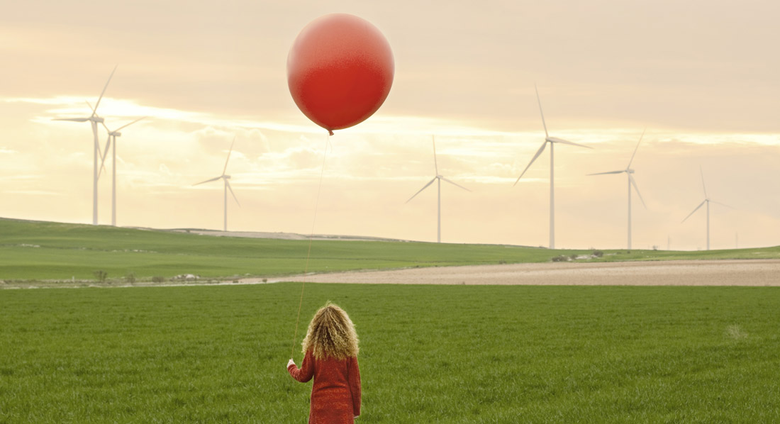 Ein Mädchen auf einer Wiese mit einem Luftballon in der Hand im Hintergrund Windräder