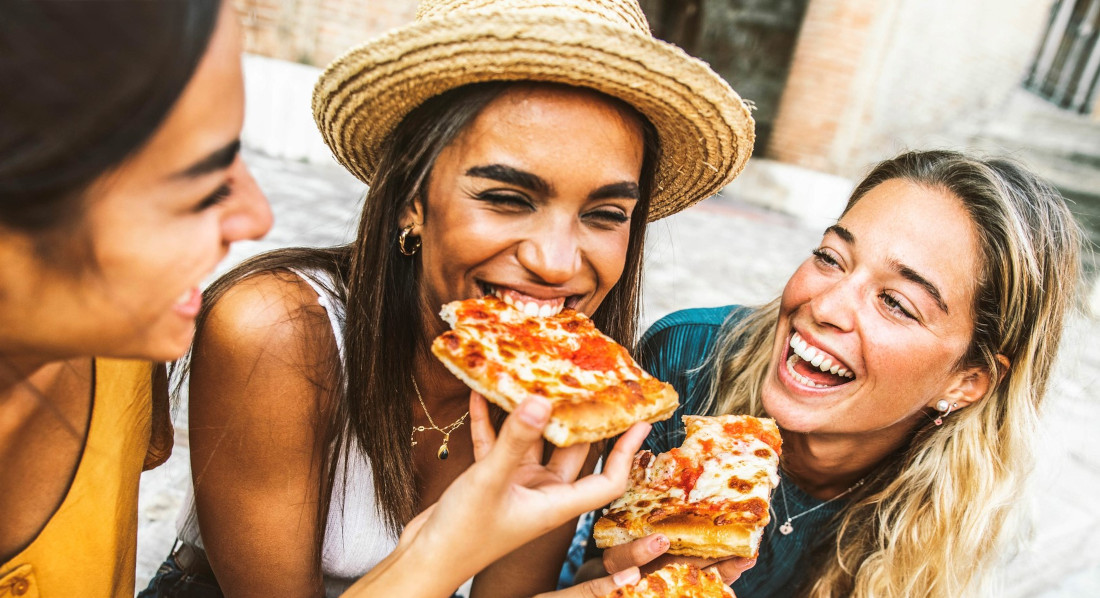 Drei junge Frauen sitzen auf einer Steintreppe und essen Pizza