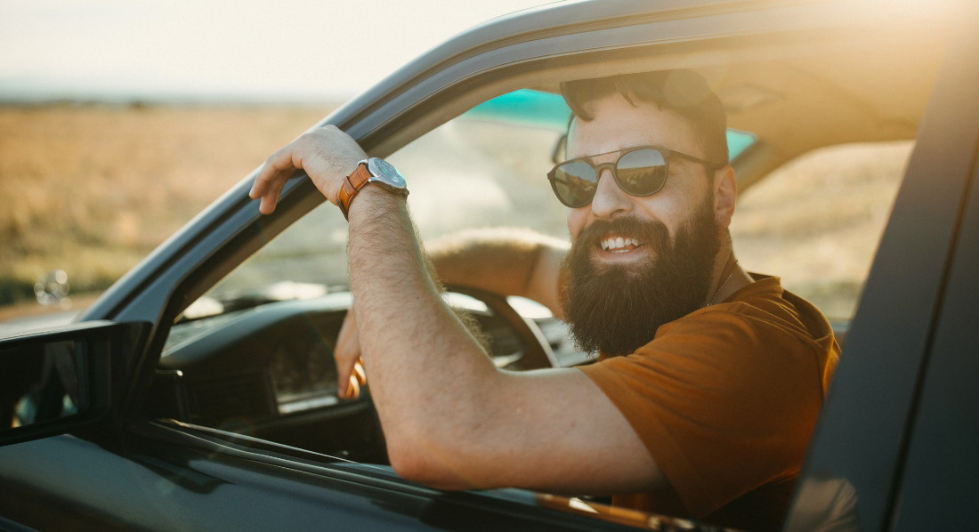 Junger Mann mit Sonnenbrille und Vollbart blickt lächelnd aus dem Autofenster