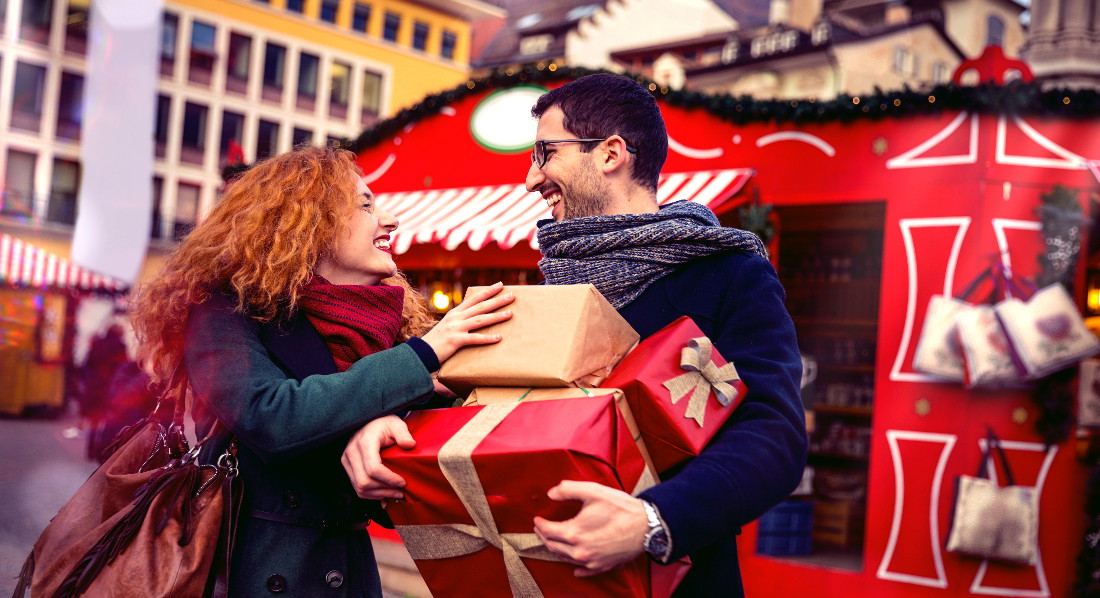 Die Frau lädt dem Mann beim weihnachtlichen Shopping mehrere Geschenkkartons auf den Arm