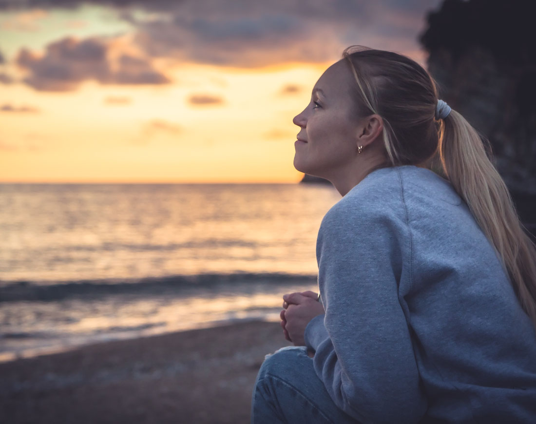 Eine junge Frau sitzt am Strand und sieht zuversichtlich dem Sonnenuntergang am Horizont zu