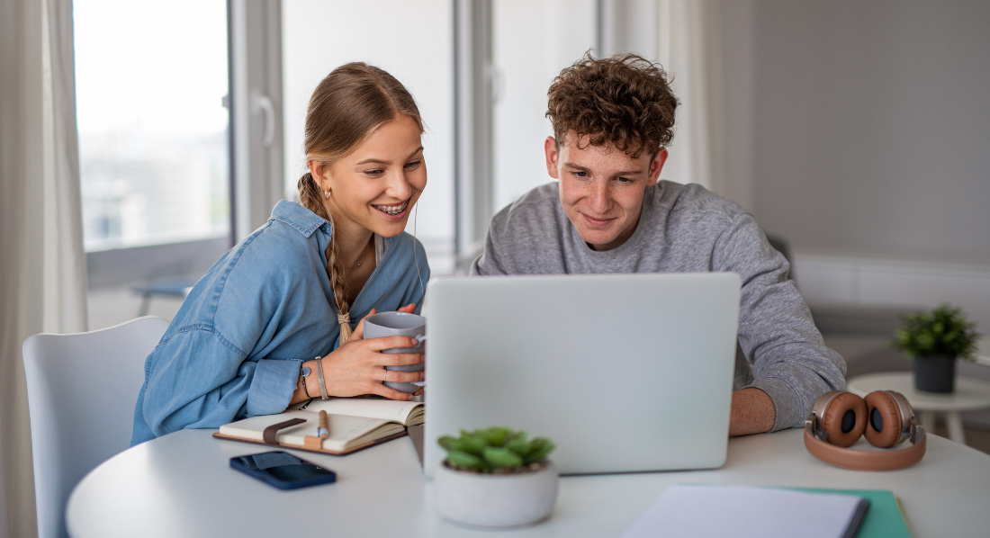 Ein junges Paar sitzt am Tisch, lächelt und schaut gemeinsam auf einen Laptop