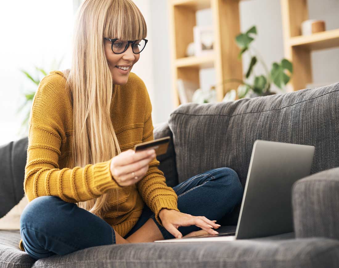 Junge Frau sitzt mit Laptop und Kreditkarte zuhause auf dem Sofa