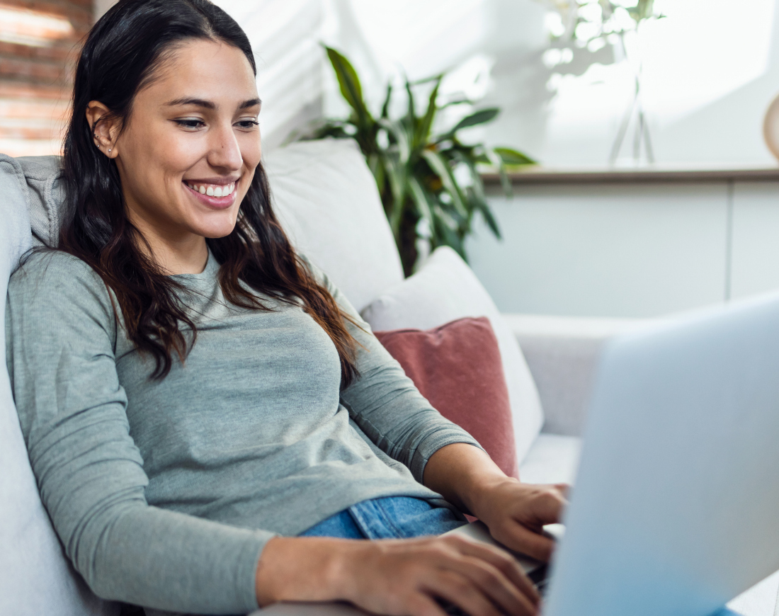 Junge Frau sitzt entspannt auf dem Sofa und tippt auf ihrem Laptop