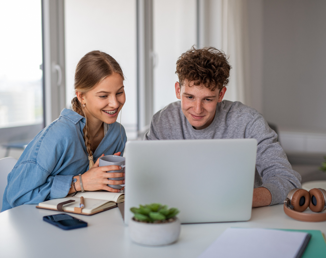 Ein junges Paar sitzt am Tisch, lächelt und schaut gemeinsam auf einen Laptop