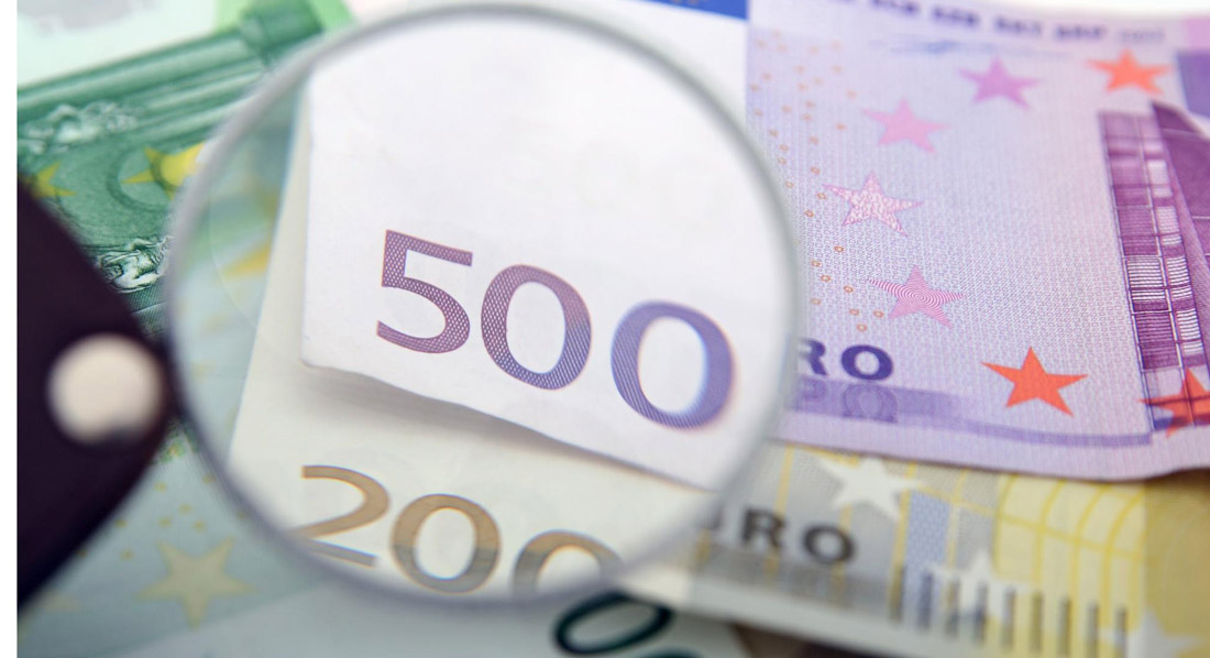 Eine Lupe vergrößert den Wert je eines 500- und 200-Euro-Scheins, die auf einem Tisch zusammen mit einem 100-Euro-Schein liegen