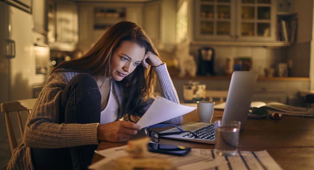 Eine gestresste junge Frau sitzt zu Hause vor ihrem Laptop und blickt verwundert auf Papiere