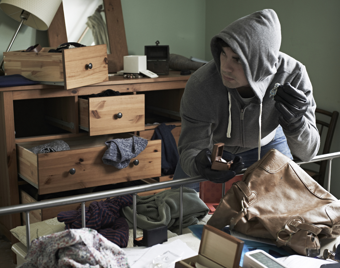 Diebstahlversicherung: Ein Einbrecher in grauer Kapuzenjacke durchwühlt ein Schlafzimmer