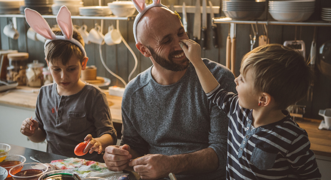 Vater bemalt mit zwei Söhnen Ostereier, Vater und ein Sohn tragen Hasenohren, ein Sohn bemalt dem Vater die Nase