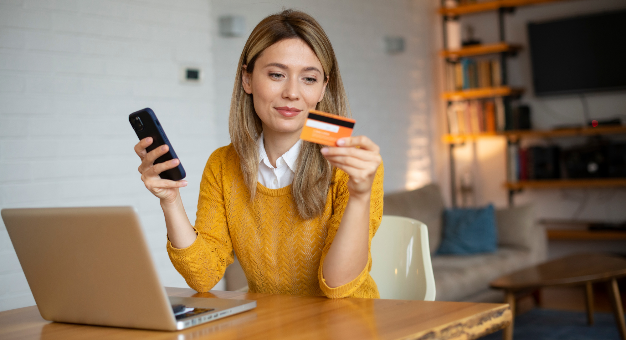 Eine Frau mit Handy und Bankkarte in den Händen führt eine SEPA-Überweisung auf einem Laptop aus