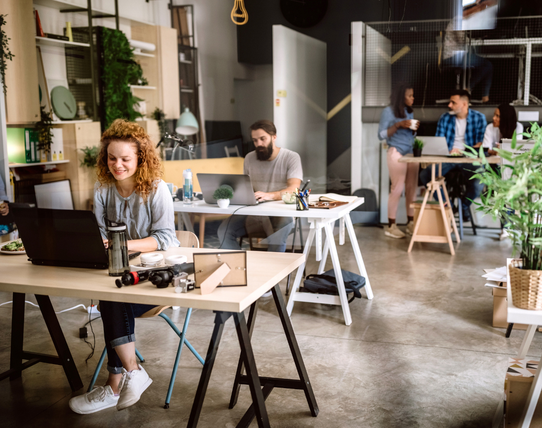 Mehrere Personen teilen sich ein Großraumbüro, sitzen mit ihren Laptops am Schreibtisch oder plaudern bei einem Kaffee