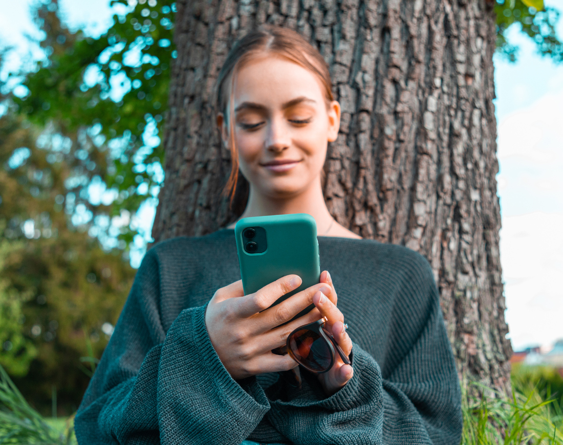 Eine junge Frau lehnt sich an einem Baum an und schaut lächelnd auf ihre CO₂-Bilanz auf einer Klima-App