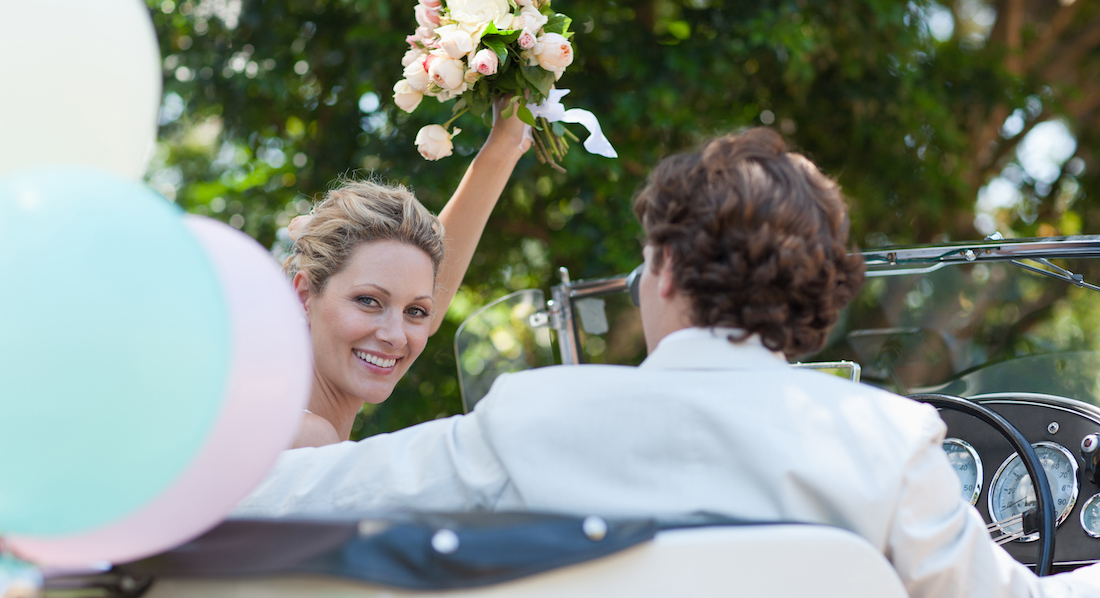 Junges Ehepaar fährt im Wagen davon, sie winkt mit dem Brautstrauß