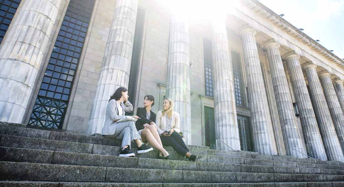 Drei junge Frauen sitzen auf der Treppe vor dem Eingangsportal einer Universität