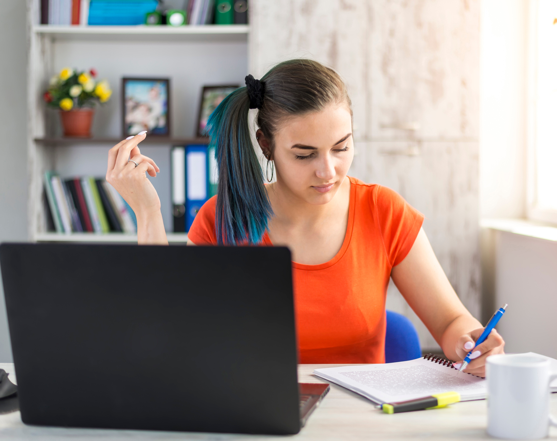 Eine jugendliche Frau sitzt am Schreibtisch vor einem Laptop und macht sich in einem Block Notizen