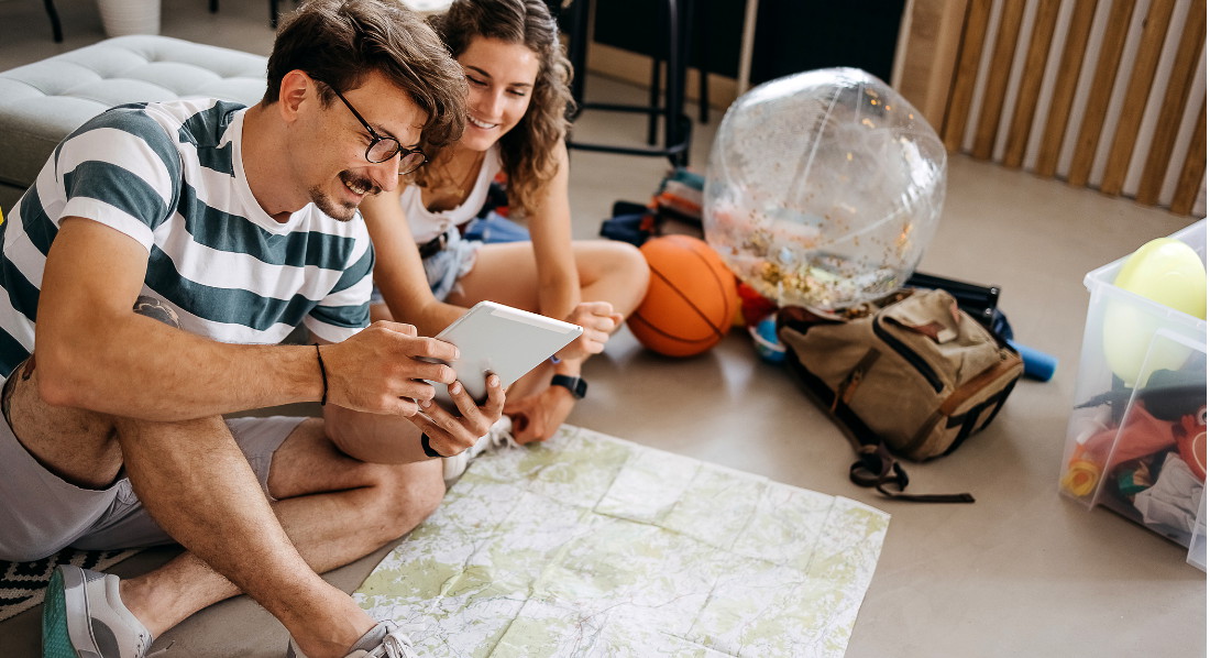 Ein junges Pärchen sitzt vor einer Mappe auf dem Boden und schaut auf ein iPad