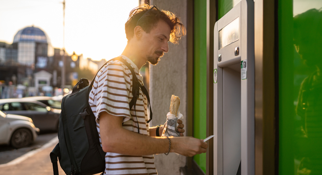Ein Mann beim Geldabheben am Automaten, im Hintergrund sind parkende Autos und Gebäude