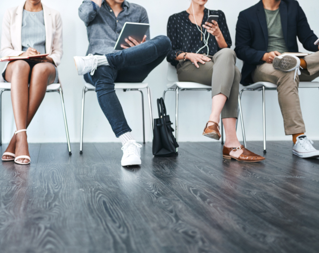 Männer und Frauen sitzen mit Laptops, Smartphone und Notizblock in einem Warteraum auf Stühlen