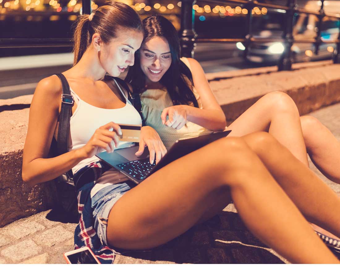 Zwei junge Frauen sitzen auf den Stufen am Rand einer nächtlichen Straße und surfen auf einem Laptop im Internet