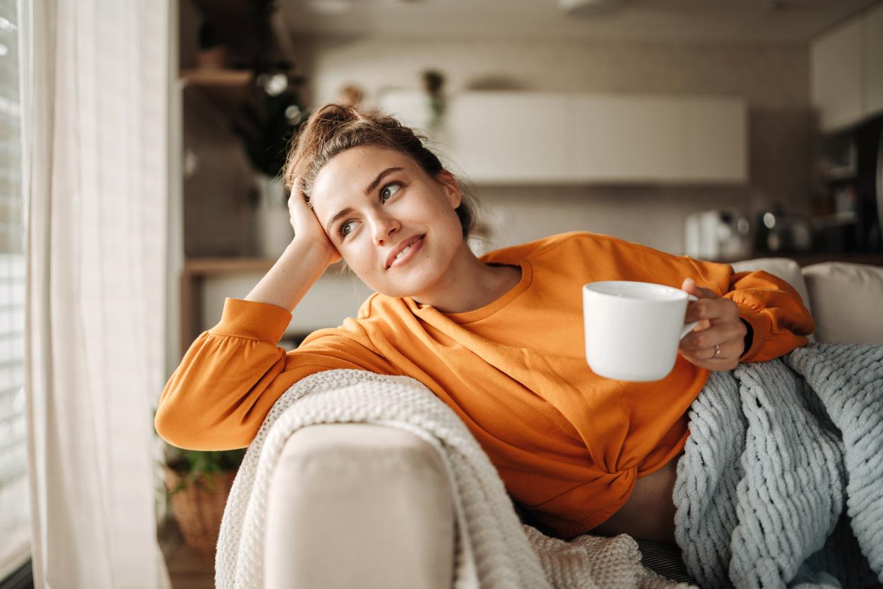 Eine Frau liegt nachdenklich auf der Couch mit einer Tasse in der Hand und denkt über die SicherAlltag Versicherung der Hanseatic Bank nach