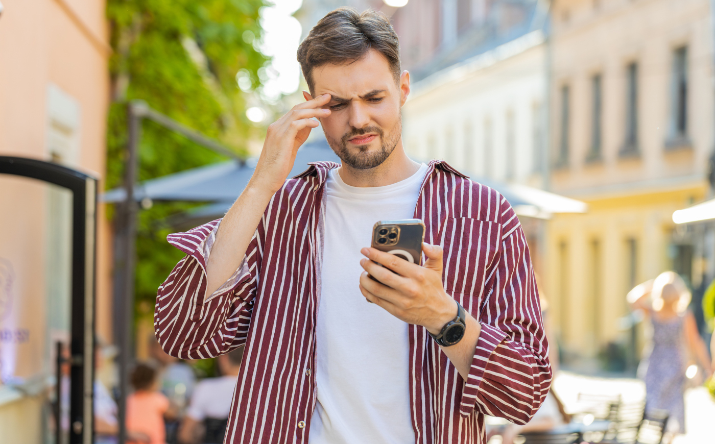 Ein verwirrt bis verärgert dreinblickender Mann mit Handy in der Hand steht in einer sonnigen, europäischen Altstadtkulisse
