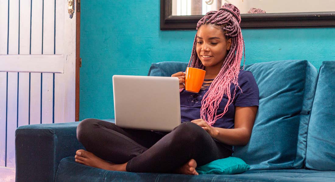 Junge Frau sitzt mit Becher und Laptop auf der Couch