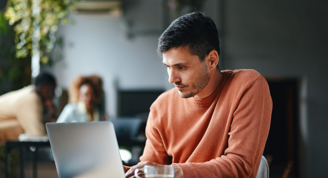Ein Mann arbeitet am Laptop an einem Schreibtisch, auf dem sich ein schwarzer Kaffee und Unterlagen befinden, im Hintergrund sind zwei weitere Menschen an einem Schreibtisch zu sehen
