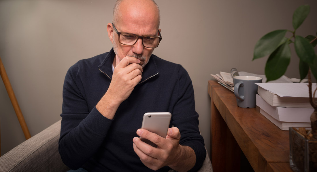 Ein Mann mittleren Alters sitzt zu Hause auf einem Stuhl und blickt kritisch auf sein Smartphonehone