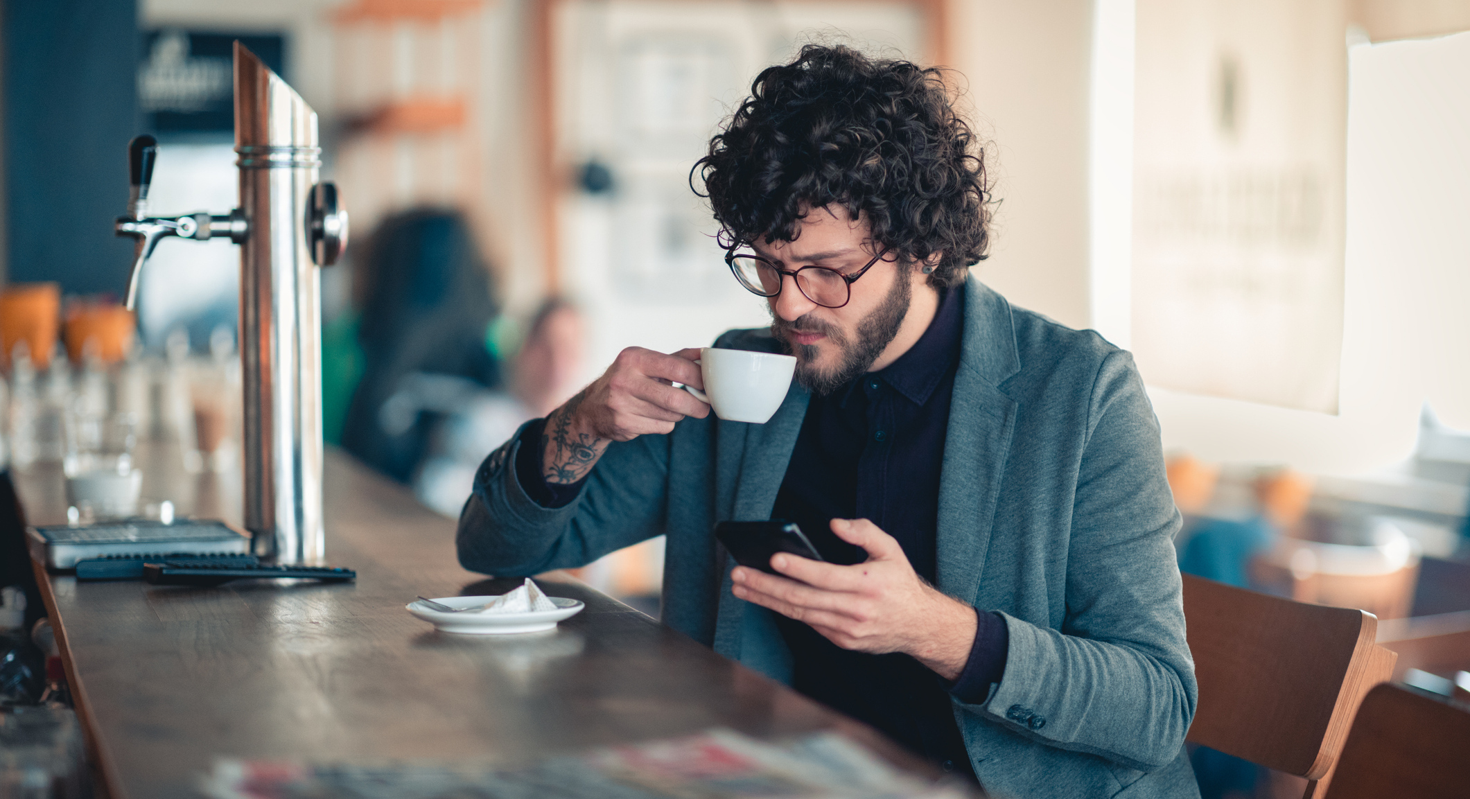 Ein bärtiger Mann sitzt am Tresen, trink Kaffee und schaut auf sein Smartphone