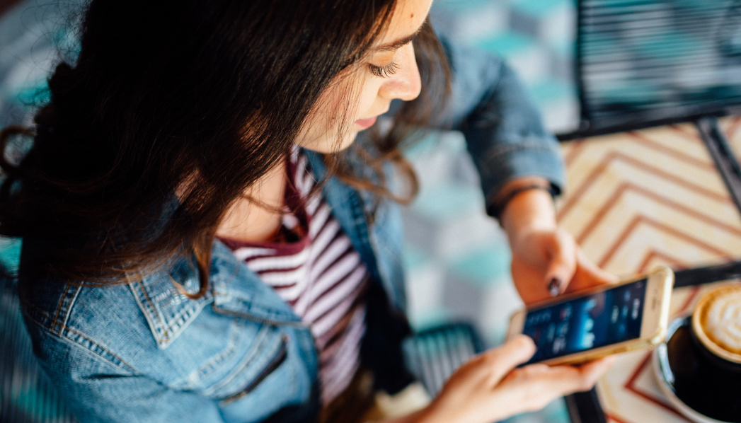 Eine junge Frau sitzt mit einem Cappuccino im Café und hält ihr Smartphone mit einer Finanz-App auf dem Display