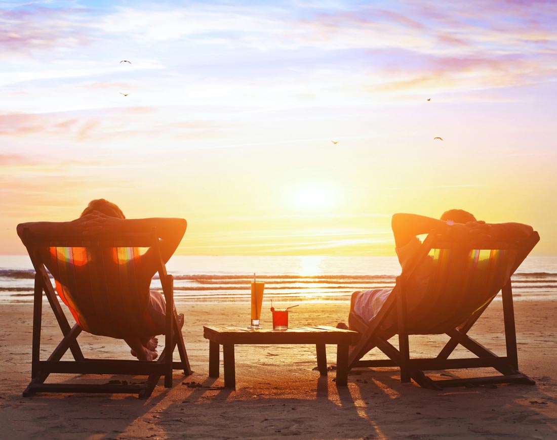 Ein Paar sitzt in Liegestühlen am Strand und verfolgt entspannt den Sonnenuntergang
