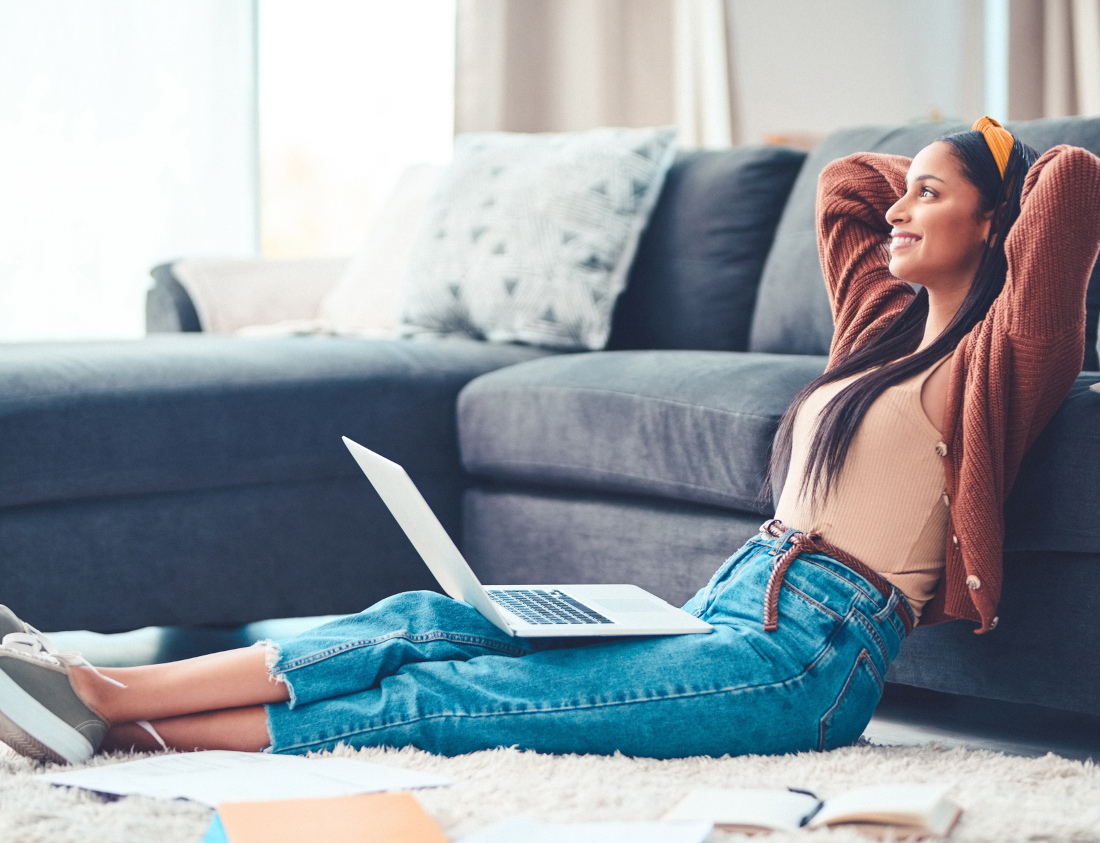 Eine Person sitzt glücklich mit ihrem Laptop und einigen Unterlagen vor ihrem Sofa auf dem Boden