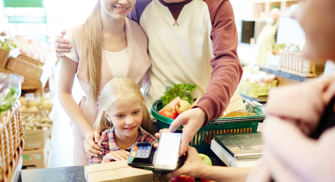 Junges Mädchen schaut zu, wie seine Eltern an der Kasse eines Gemüsemarkts per NFC bezahlen