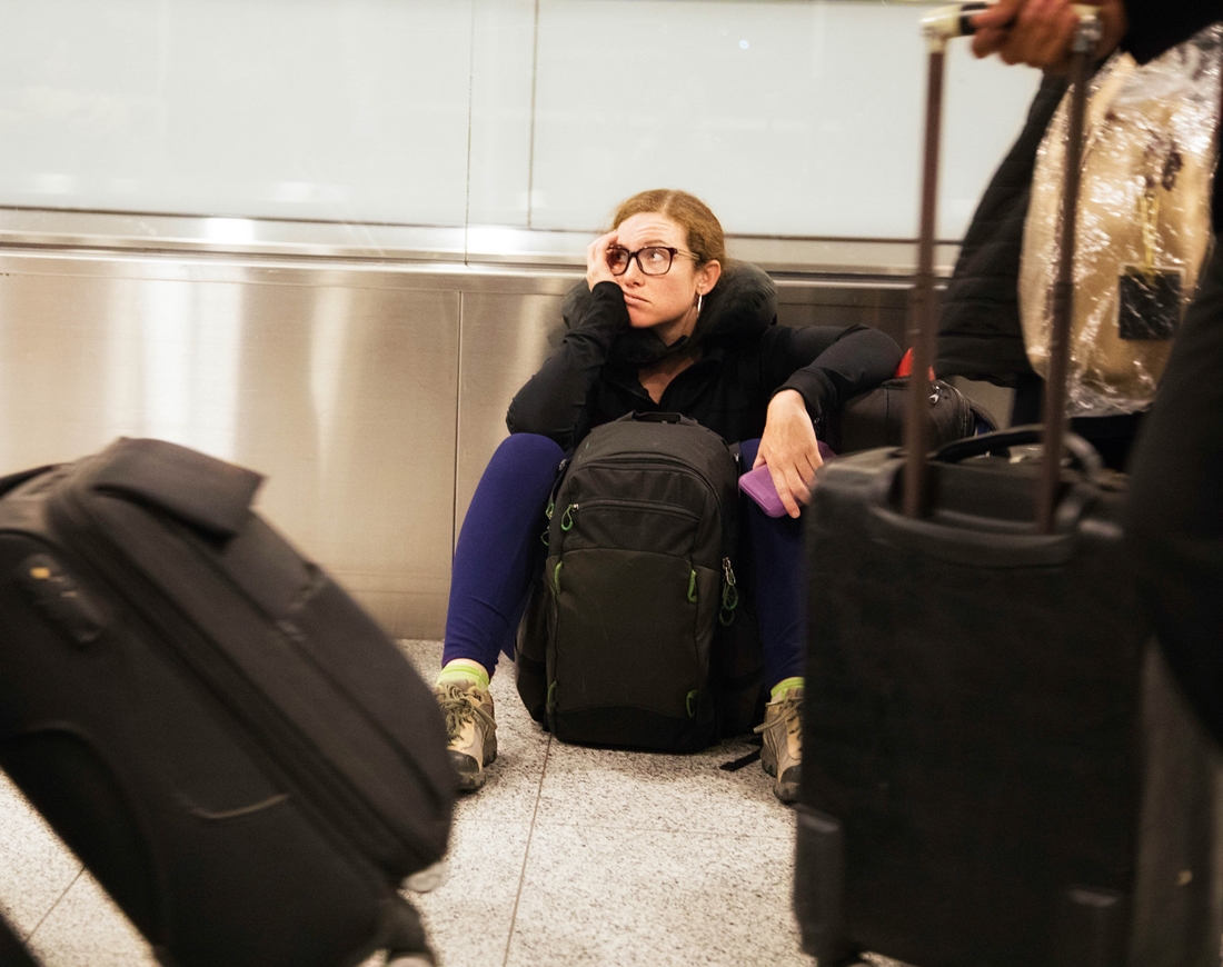 Junge Frau sitzt mit ihrem Rucksack genervt auf dem Boden am Flughafen