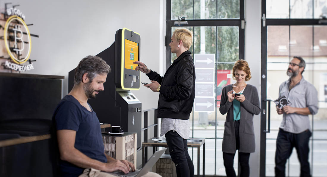 Eine Person steht mit Smartphone an einem Bitcoin-Automaten, hinter ihr warten Leute