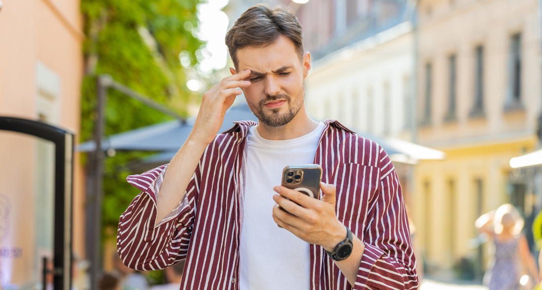 Ein verwirrt bis verärgert dreinblickender Mann mit Handy in der Hand steht in einer sonnigen, europäischen Altstadtkulisse