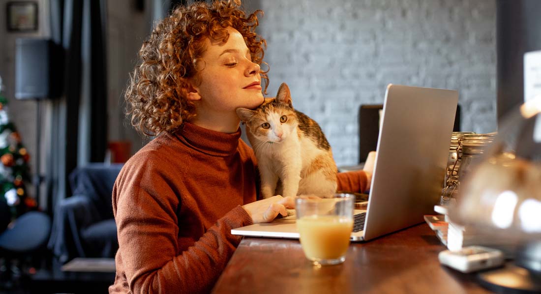 Eine junge Frau arbeitet an ihrem Laptop, während sich ihre Katze an ihr Kinn schmiegt