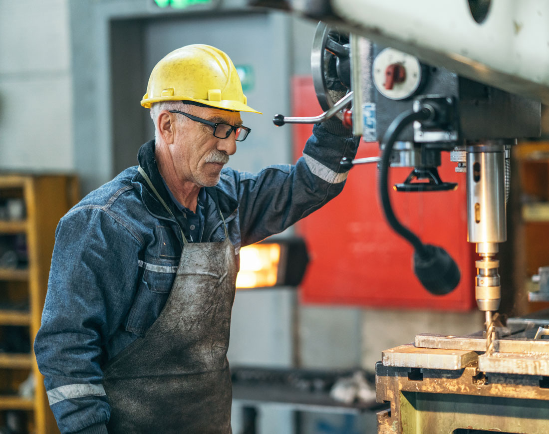 Älterer Arbeitnehmer mit Schutzhelm steht an einer Werkbank