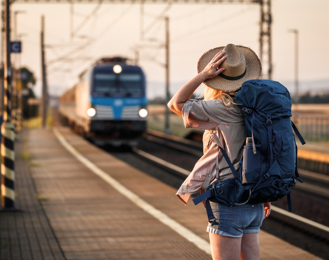 Frau mit Backpacking-Rucksack und Hut steht am Bahngleis und blickt einfahrendem Zug entgegen