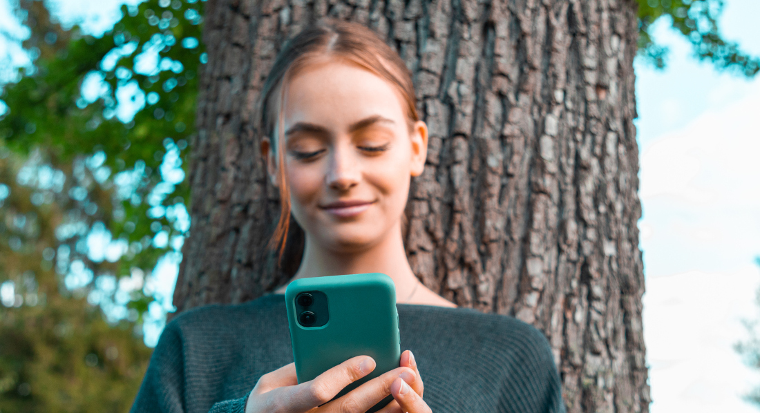 Eine junge Frau lehnt sich an einem Baum an und schaut lächelnd auf ihre CO₂-Bilanz auf einer Klima-App