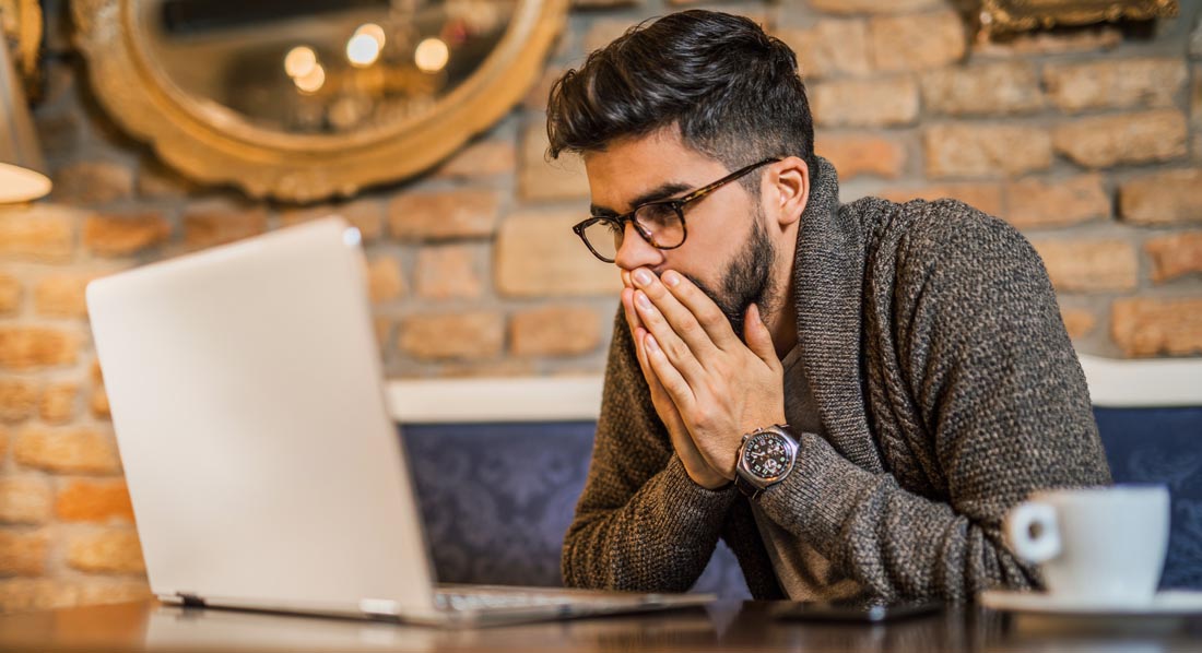 Schockiert wirkender junger Mann vor einem Laptop hält sich die Hände vor den Mund