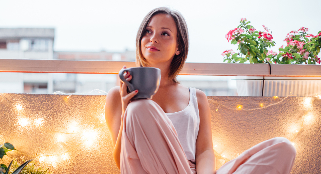 Eine junge Frau in Freizeitkleidung sitzt entspannt mit einem großen Becher in der Hand auf dem Boden eines Balkons