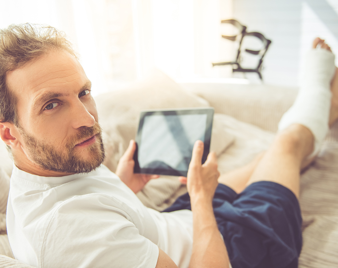 Ein junger Mann liegt mit einem Gipsbein auf einem Sofa und hält ein Tablet in der Hand