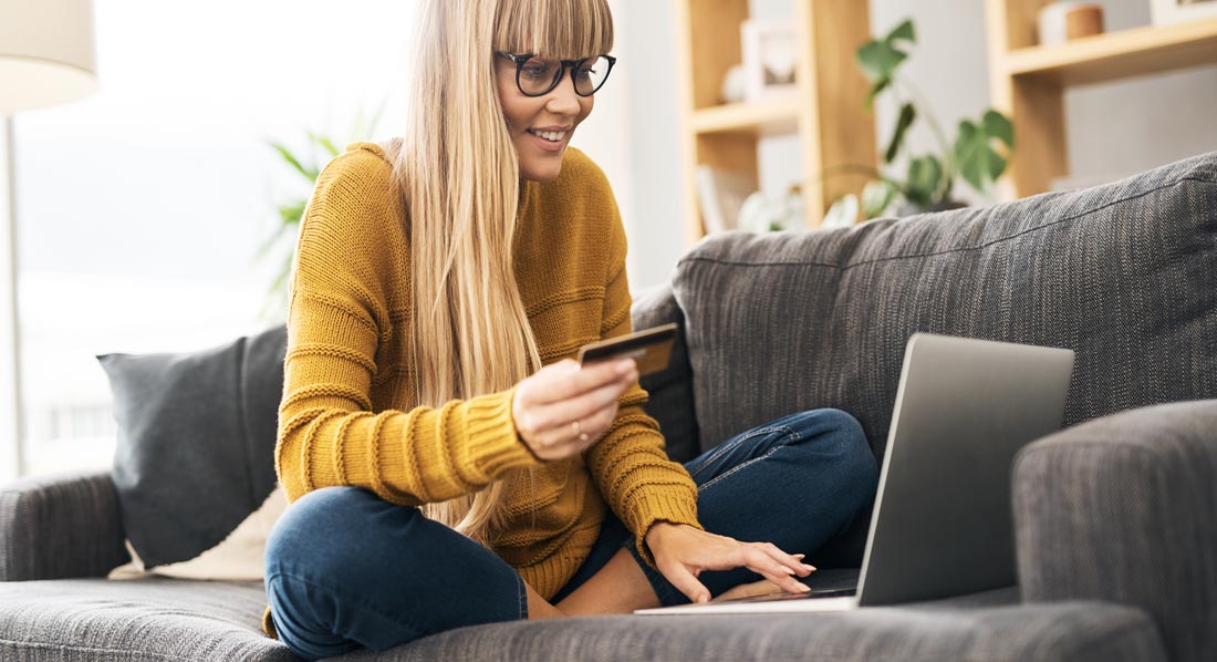 Junge Frau sitzt mit Laptop und Kreditkarte zuhause auf dem Sofa
