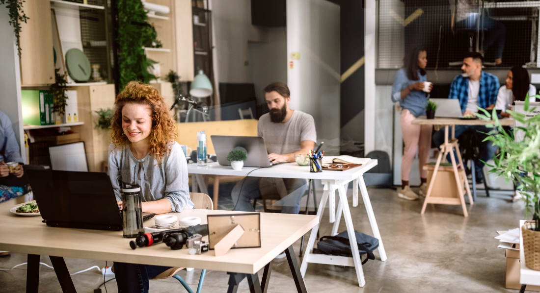 Mehrere Personen teilen sich ein Großraumbüro, sitzen mit ihren Laptops am Schreibtisch oder plaudern bei einem Kaffee