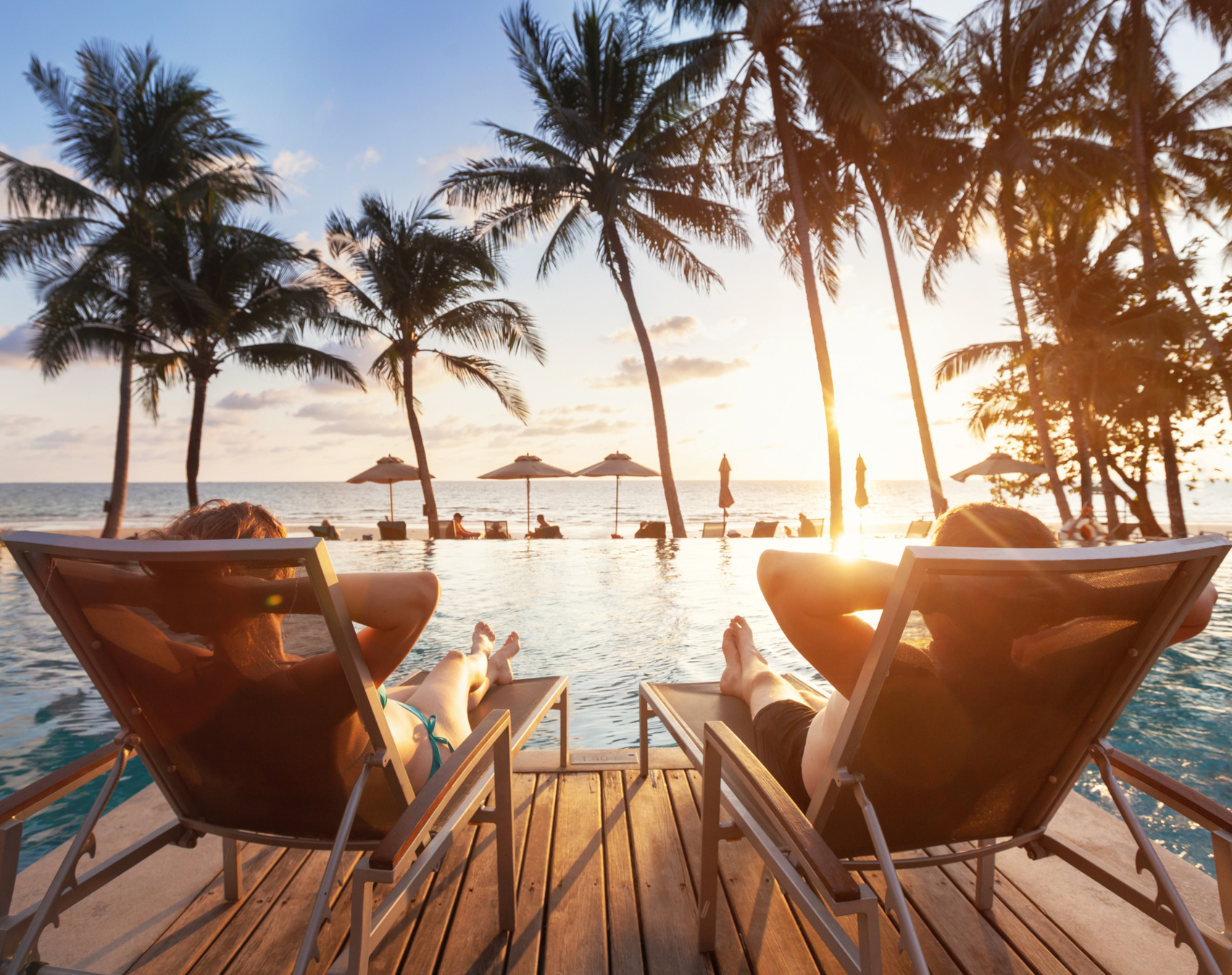 Zwei Personen auf Liegestühlen vor einem Pool und Palmen sehen in den Sonnenuntergang