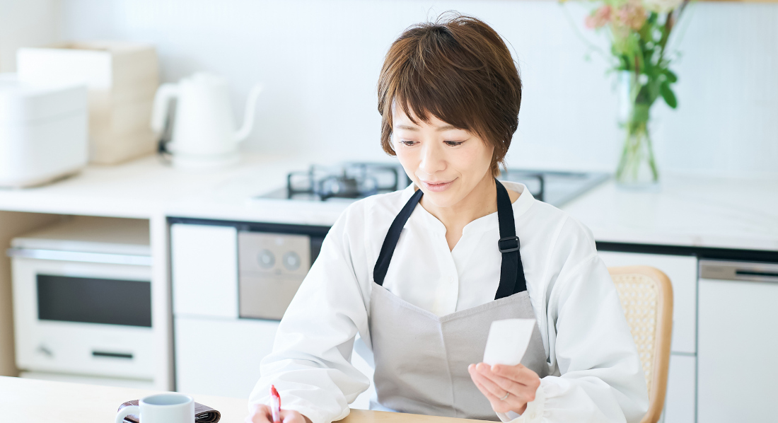 Eine Frau sitzt am Küchentisch, vor sich ein aufgeschlagenes Notizbuch, und prüft Quittungen.